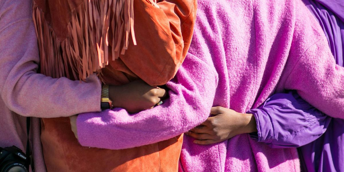 Close up of four women standing arm in arm with their backs to the camera. They are wearing bright orange, pink and purple coats