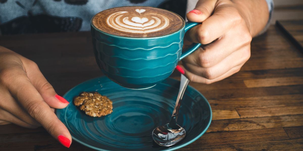 woman's hands, with red nail polish, hold a cappuccino in a turquoise cup over a saucer with a small, oaty biscuit on one side and a teaspoon on the other