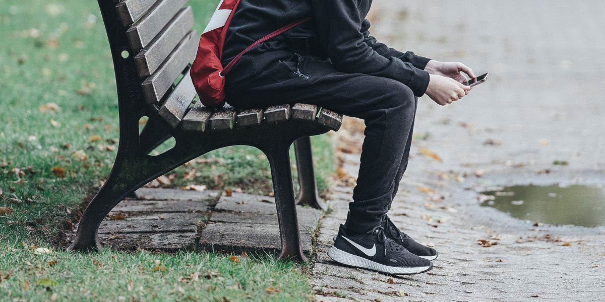 A child in a black tracksuit and red backpack sits on a park bench holding a mobile phone