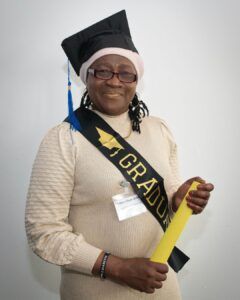 Woman in cream jumper dress, graduation hat and sash, holds her graduation scroll