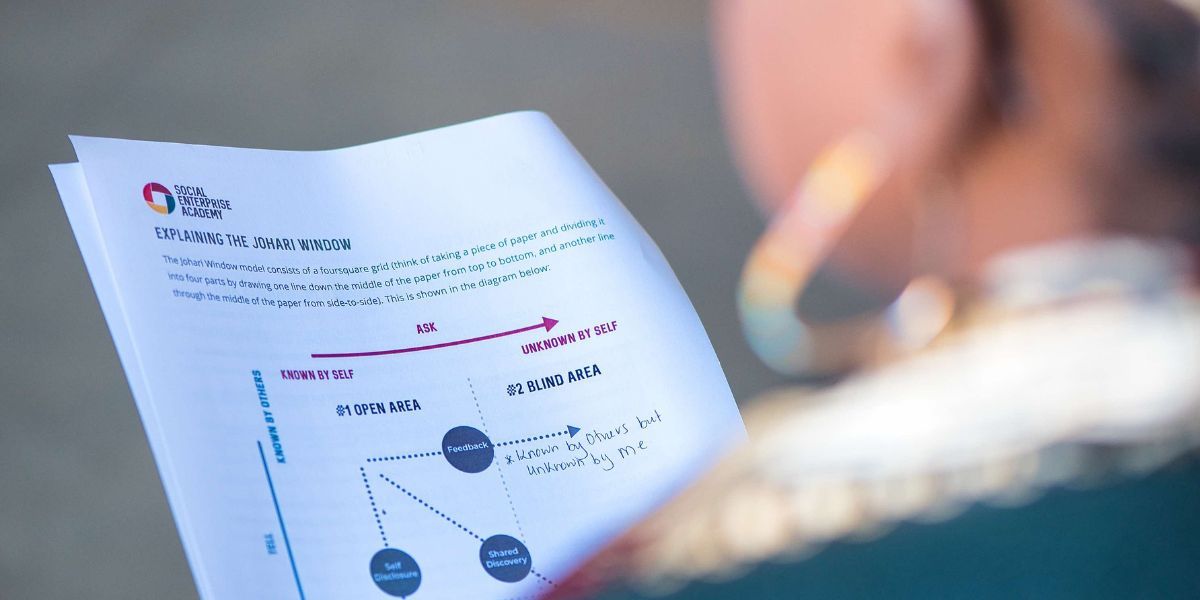 Peering over the left shoulder of a woman wearing a white shirt, green jumper and large gold hopped earring who appears in soft focus. She is reading an exercise sheet containing graphs and text, with a Social Enterprise Academy logo in the top left hand corner.