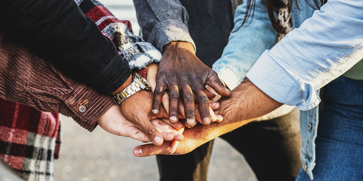 Six hands of people from different ethnic backgrounds meet in the middle of the image in a show of community