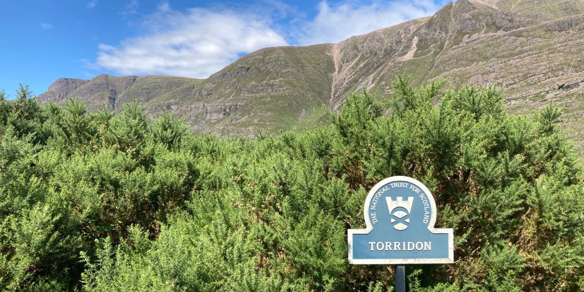 Torridon sign post with greenery, Liathach mountain and a blue cloudy sky in the background