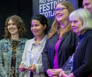 a diverse group of smiling women stand in front of a black Refugee Festival Scotland banner, some holding glass awards