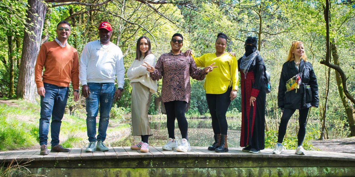 Seven smiling people (two men and five women) stand side by side on a bridge in a leafy, green forest