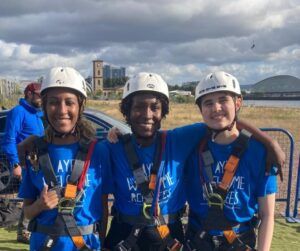 Three women in blue Scottish Refugee Council tshirts, harnesses and white hard hats smile for the camera as a person zipslides across the Glasgow skyline in the background