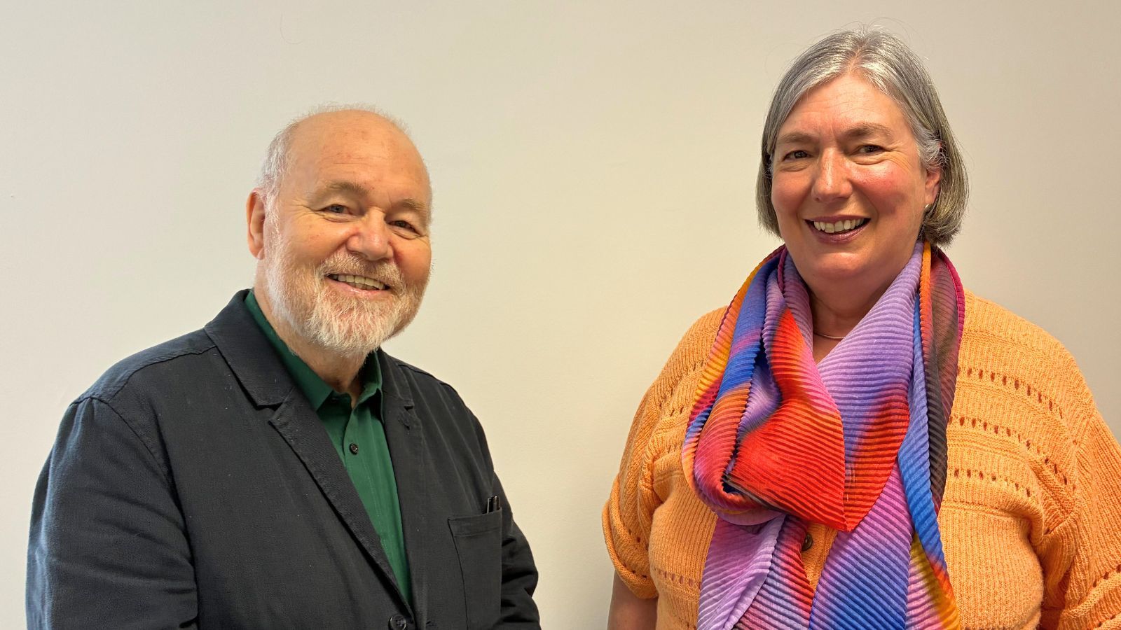 A smiling grey haired man with a beard poses alongside a smiling grey aired women wearing a colourful scarf