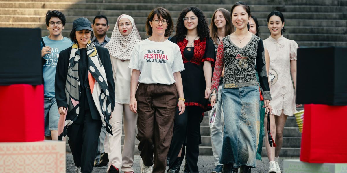 Nine happy, smiling people walk towards the camera in a line. They are wearing brightly coloured clothes. One has a t-short that reads 'aye welcome refugees' the person in the centre wears a t-shirt that reads 'Refugee Festival Scotland'