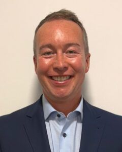 Colin Macfarlane - smiling close up of a man with graying hair wearing a white shirt wityh top button undone and a blue suit jacket 