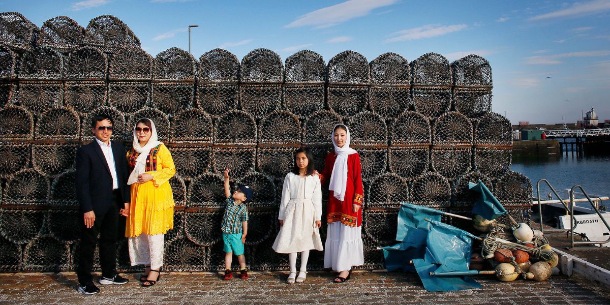 Smiling family of five in colourful Afghan clothing pose in front of a wall of lobster pots by the shore. The image is one of a collection of New Scots Portraits by award-winning photojournalist Angela Catlin.