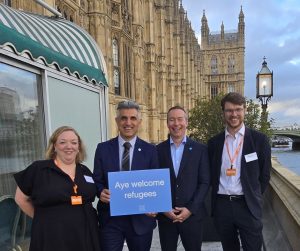 Three smiling men and one woman in smart clothes pose for a photograph in front of Westminster holding a blue placard with a Scottish Refugee Council logo and white writing that reads 'aye welcome refugees'